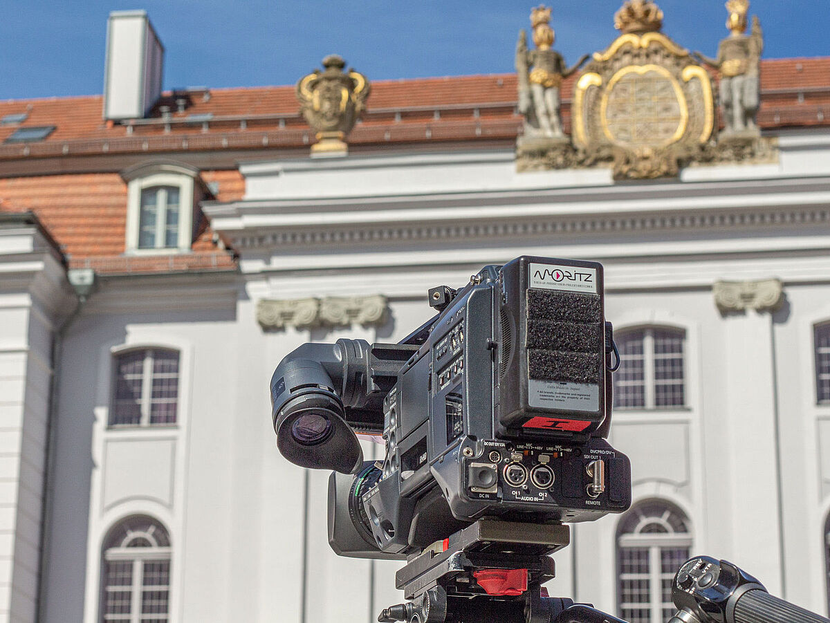 Symbolbild Wissenschaftskommunikation Im Vordergrund ist eine professionelle Filmkamera auf einem Stativ zu sehen, im Hintergrund das Unihauptgebäude mit der weißen Barockfassade mit verzierten Elementen und roten Dachziegeln.